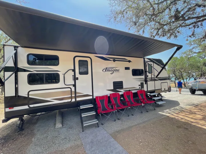This Imagine RV rental shows a comfy outdoor setup: an extended awning shades red chairs at a gravel campsite.. Grand Design Imagine 2021