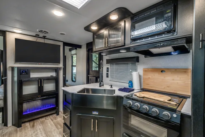 Modern RV rental interior: dark wood kitchen with sink, stove, and microwave, plus living area with TV and electric fireplace.. Cherokee Grey Wolf 2022