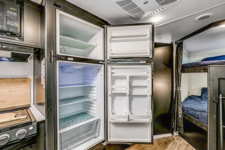 RV rental interior showcasing an open two-door refrigerator, microwave, stove, and adjacent bunk beds with blue bedding.. Cherokee Grey Wolf 2022