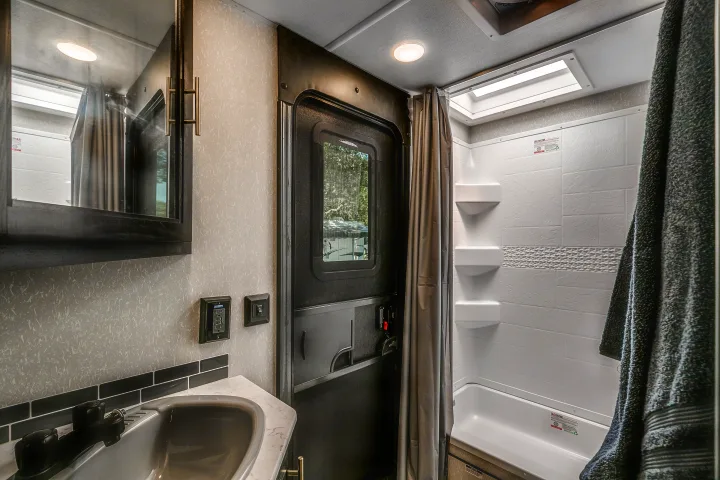 RV rental bathroom with a compact sink, mirror, and white shower. A dark door and gray shower curtain are visible.. Cherokee Grey Wolf 2022