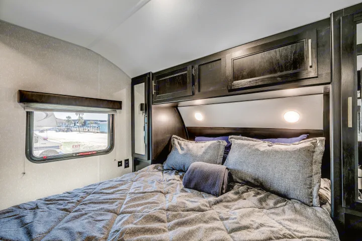A modern RV rental bedroom featuring practical dark wood cabinetry, soft grey bedding, and a view of the campground.. Cherokee 26DBH 2022