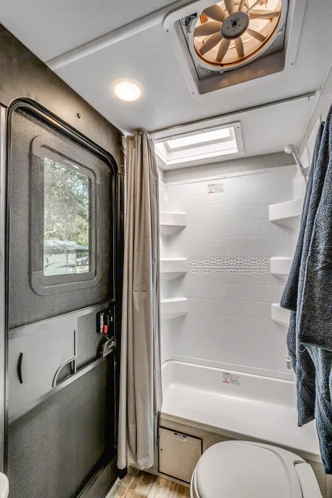 Inside an RV rental bathroom, showing a shower-tub, toilet, ceiling fan, skylight, and the main entry door.. Cherokee 26DBH 2022