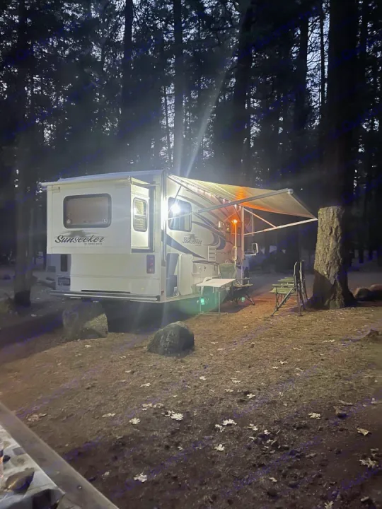 Illuminated RV rental under a dark forest sky. Awning, chairs, and table set for a cozy night at the campsite.. Forest River Sunseeker LE 2014