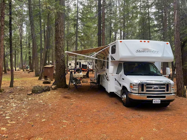 White Sunseeker RV rental parked at a forest campsite. Awning extended, people enjoying the outdoor setup among tall trees.. Forest River Sunseeker LE 2014