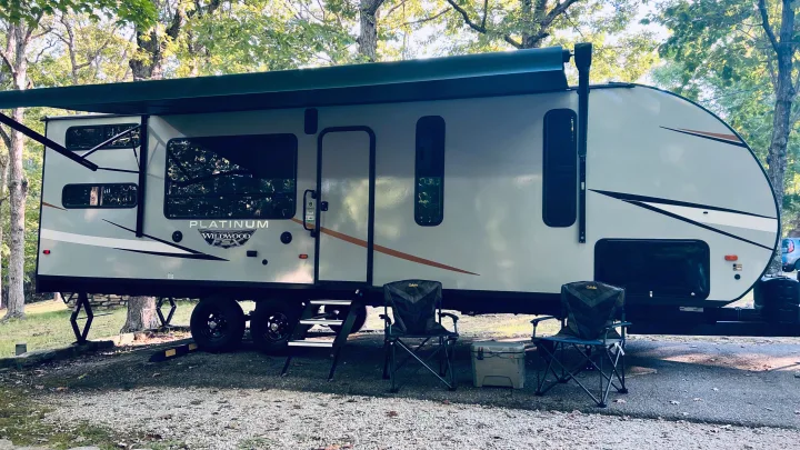 This RV rental is set up at a leafy campsite with chairs and a cooler, ready for outdoor adventures.. Forest River Wildwood 2022