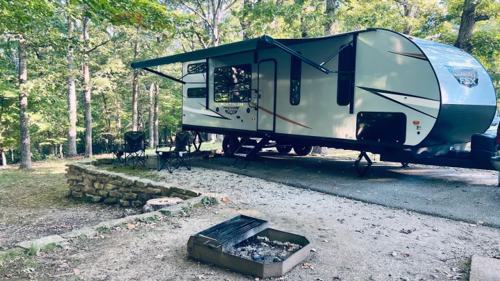 RV rental parked at a spacious gravel camping site among trees, complete with an open awning, chairs, and a fire pit.. Forest River Wildwood 2022