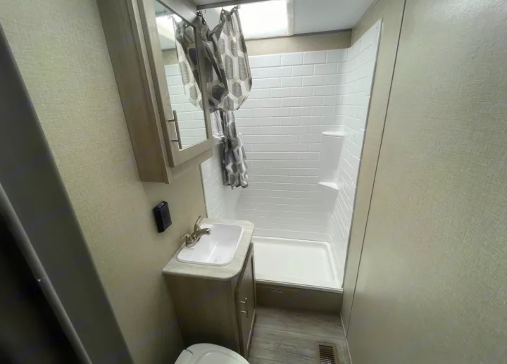 This RV rental bathroom features a compact sink, mirror, shower with a gray patterned curtain, and toilet, maximizing space for travel.. Forest River Coachmen Catalina Legacy 2022