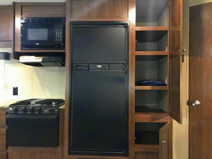 Compact RV rental kitchen with black refrigerator, microwave, stove, and built-in pantry/storage in wood cabinetry.. Dutchmen Coleman Lantern 2017