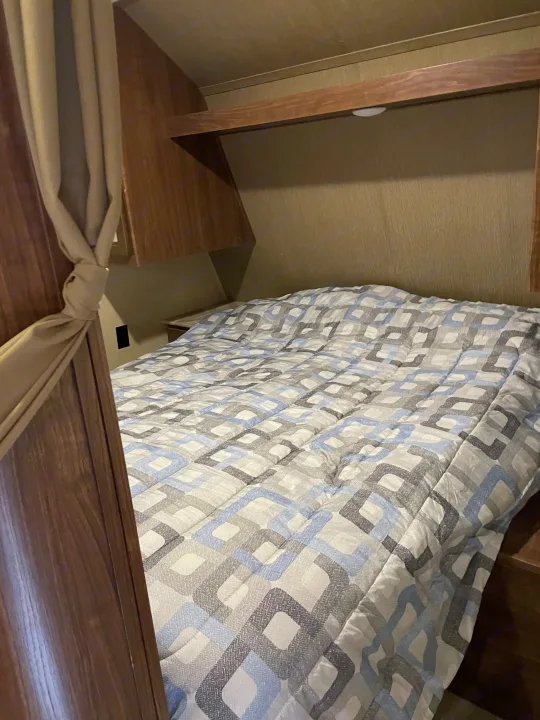 Cozy bed inside an RV rental, featuring a blue and grey patterned comforter, wooden walls, and a tied-back curtain.. Dutchmen Coleman Lantern 2017