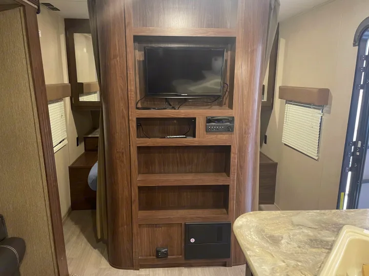 RV rental interior: A central wooden entertainment unit with TV, stereo, and shelving, adjacent to a sleeping area and kitchen space.. Dutchmen Coleman Lantern 2017