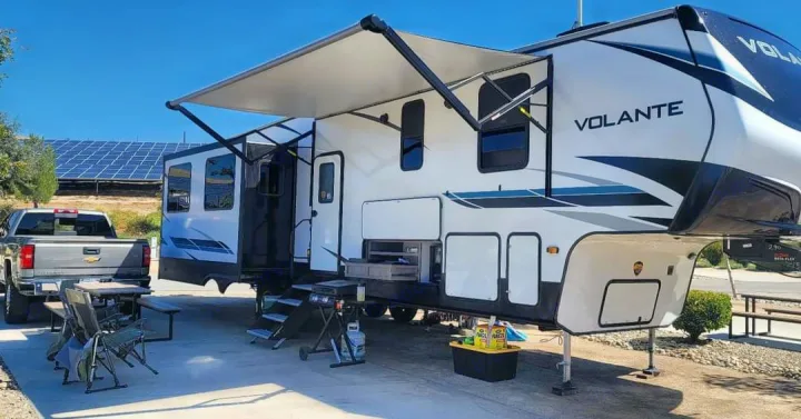 An RV rental with an extended awning, grill, and truck at a sunny campsite. Solar panels line the distant hill.. Crossroads RV Volante HP 2022
