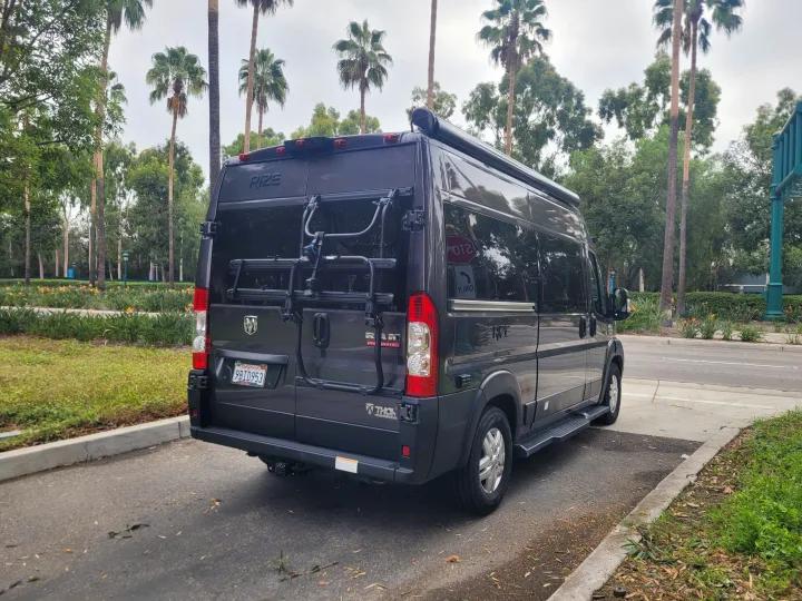 A dark Thor Motor Coach Rize camper van RV rental with a bike rack is parked amidst lush greenery and palm trees.. Thor Motor Coach Rize 18M 2023