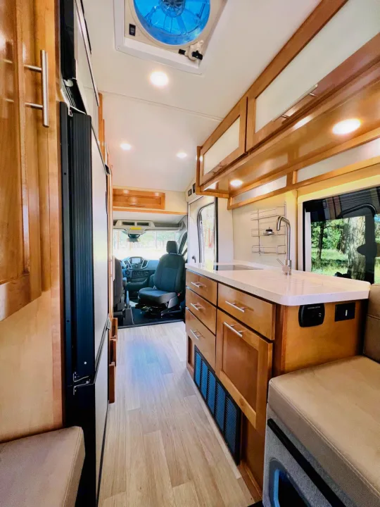 This modern rv rental interior features a compact kitchen with wood cabinets, sink, fridge, and seating, leading to the front cabin.. Coachmen Beyond 22D 2020