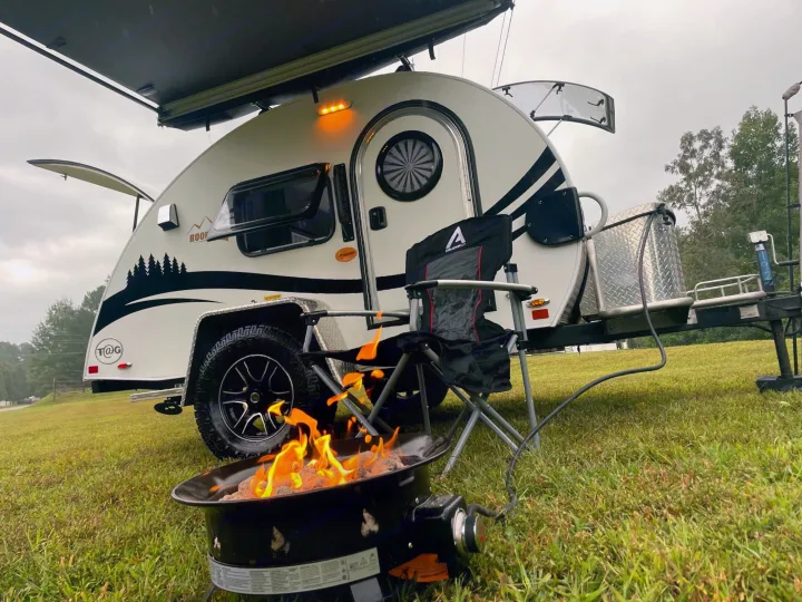 An RV rental teardrop camper with an awning extends over a cozy camp setup, featuring a portable fire pit and chair.. nuCamp T@G 2021