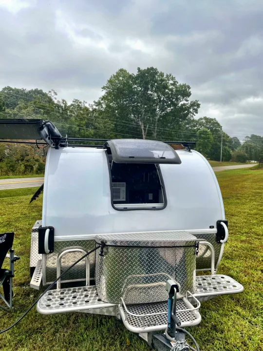 Experience outdoor freedom with this compact teardrop RV rental, parked on grass with an open window and awning on a cloudy day.. nuCamp T@G 2021
