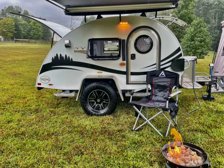 A white teardrop RV rental is set up for camping, with an outdoor chair and a glowing fire pit on green grass.. nuCamp T@G 2021