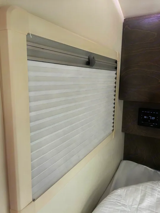 Inside an RV rental: a pleated window blind, dark wood cabinet, digital display, and part of a bed with white bedding.. nuCamp T@G 2021
