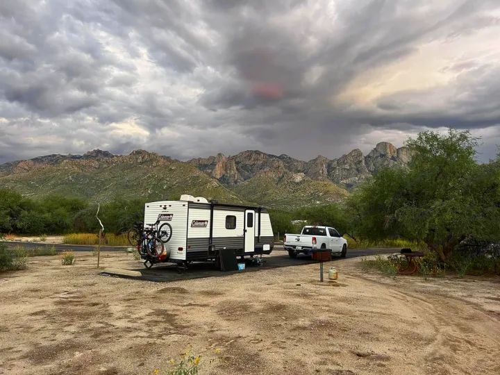 White RV rental, pickup truck, and bikes at a scenic mountain campsite under a dramatic, cloudy sky.. Coleman Other 2021