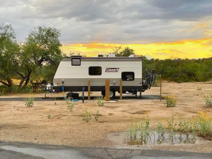 A Coleman RV rental is parked at a serene desert campsite, bathed in the glow of an incredible orange sunset behind dramatic storm clouds.. Coleman Other 2021