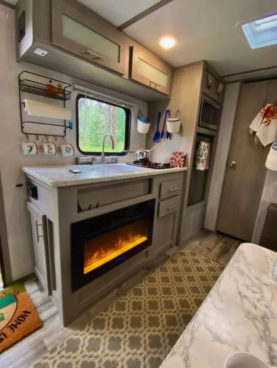 This cozy RV rental kitchen features a sink, microwave, cabinets, and a warm electric fireplace, with a window to nature. Perfect for your road trip!. Coleman Other 2021