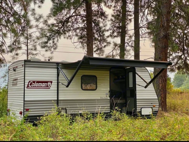 A Coleman travel trailer RV rental with an extended awning is nestled among pine trees in a grassy area.. Coleman Other 2021