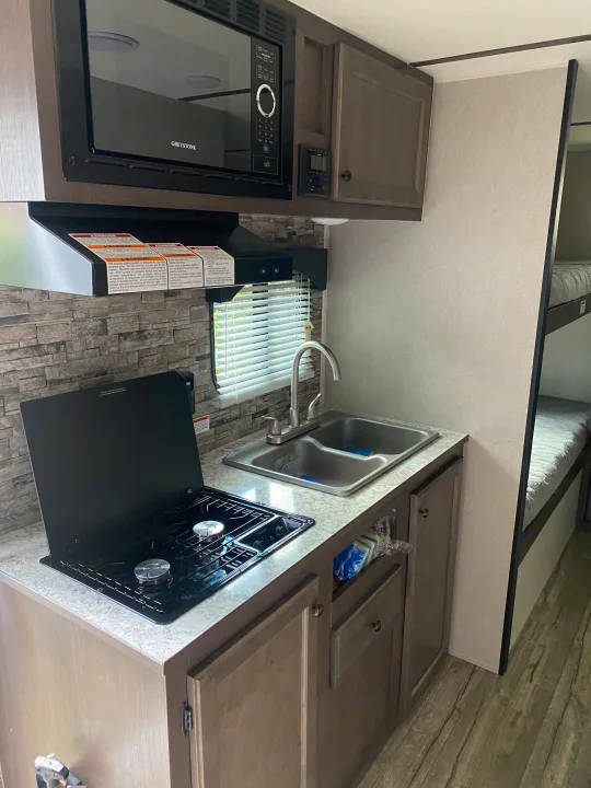 This RV rental features a compact kitchen with a microwave, stovetop, sink, and nearby bunk beds for comfortable travel.. Sunset Park RV Sun Lite 2020
