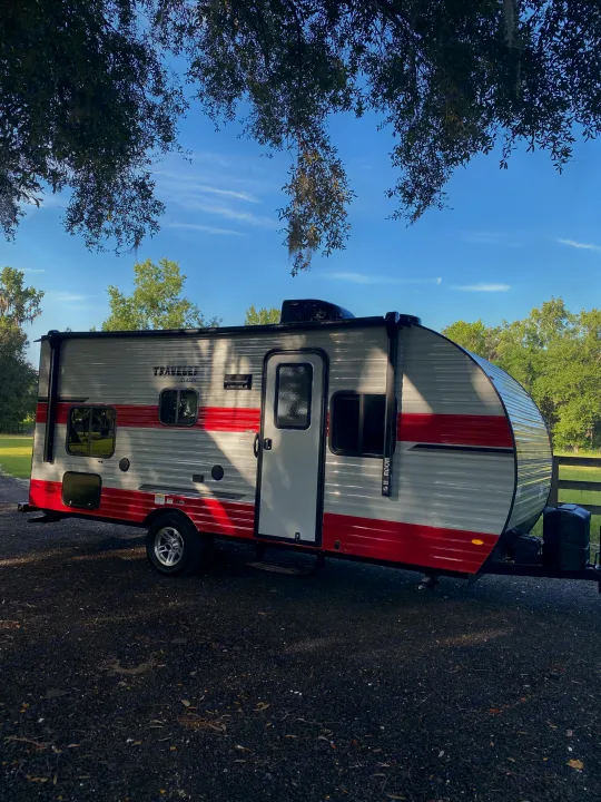 This striking gray and red travel trailer, ideal for an RV rental, is parked on gravel under shady trees.. Sunset Park RV Sun Lite 2020