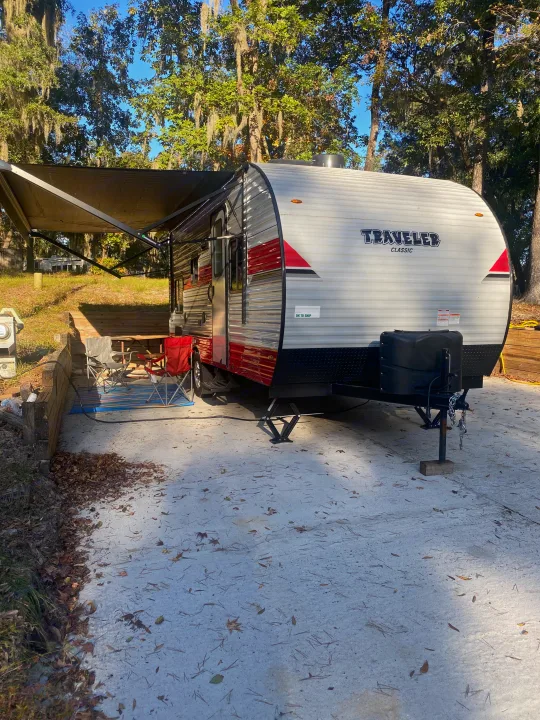 Traveler Classic rv rental parked at a sunny campsite, with awning extended and chairs set up, surrounded by mossy oak trees.. Sunset Park RV Sun Lite 2020