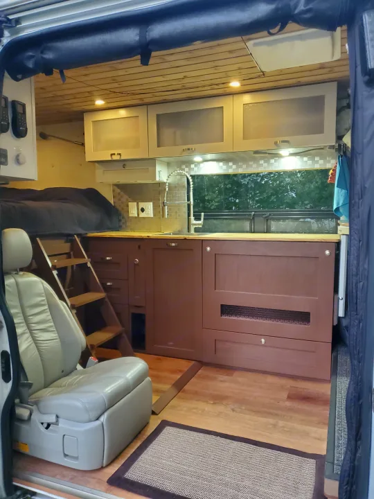 RV rental interior: Elevated bed with ladder, full kitchenette, and a comfortable swiveling seat in a cozy camper van.. Custom Camper 2017