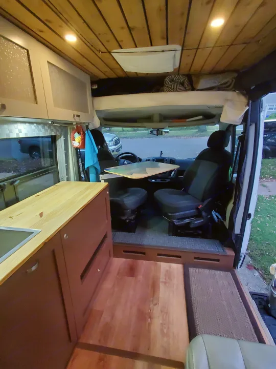 Interior of a custom camper van conversion for RV rental. Features a kitchenette, front seats with a table, and elevated sleeping area.. Custom Camper 2017
