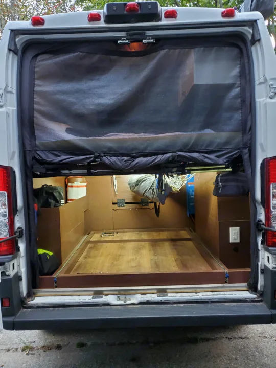 Open rear of an RV rental camper van, revealing its custom wood interior with storage, bed slide-out, and mesh screen.. Custom Camper 2017