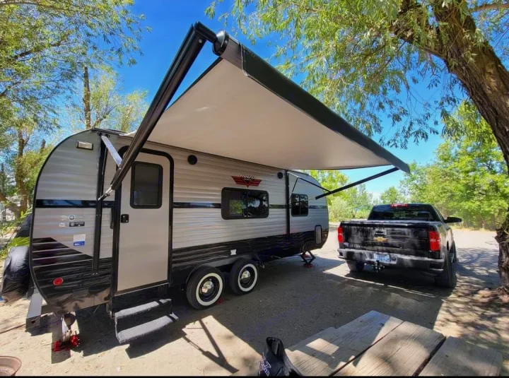 This RV rental, a Retro travel trailer with an extended awning, is parked next to a black pickup truck under trees.. Riverside RV Retro 2019