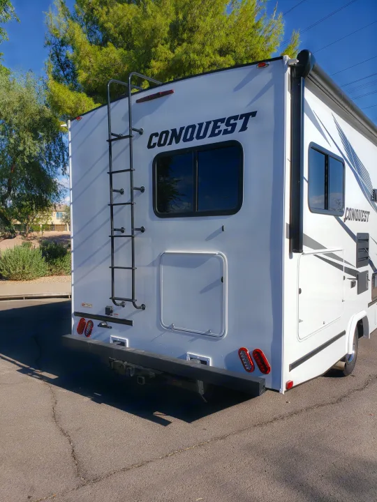 Rear and side of a white Conquest RV, great for rv rental. Features a ladder, windows, storage, and taillights. Parked outdoors.. Gulf Stream Conquest 2022