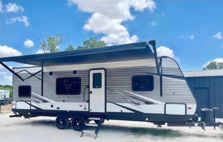 Modern grey & white travel trailer RV rental with extended awning and steps, parked outdoors under a blue sky.. Heartland RVs Trail Runner 2020
