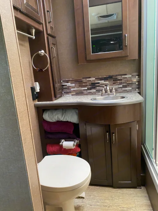 RV rental bathroom features a sink, toilet, mirrored cabinet, and open shelving with neatly folded towels, all in a compact space.. Thor Hurricane 2018