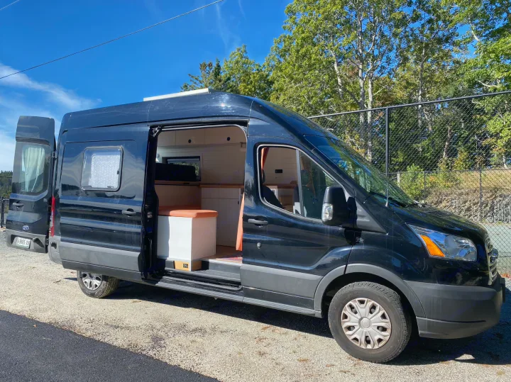 Black camper van RV rental with open side and rear doors, revealing its custom wood and white interior on a sunny day.. Ford Transit 2019
