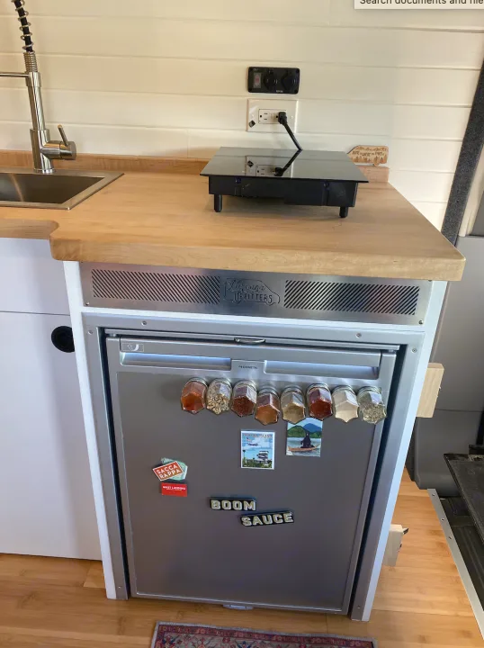 Compact RV rental kitchen with sink, portable cooktop, and fridge. Magnetic spices and fun magnets add charm to this functional space.. Ford Transit 2019