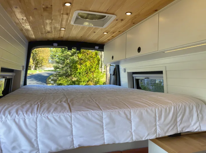 Cozy RV rental van interior featuring a comfortable bed, warm wood ceiling, and open back doors revealing a scenic outdoor view.. Ford Transit 2019