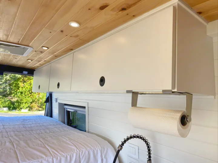 Inside an RV rental: Wooden ceiling lights illuminate white overhead cabinets, a bed, and a paper towel dispenser in the compact space.. Ford Transit 2019