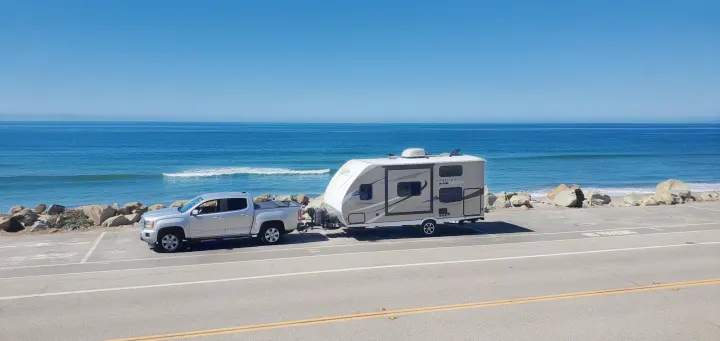 Silver pickup truck with RV rental parked by a rocky coast, facing the blue ocean. Clear sky, distant land.. Coachmen Other 2019