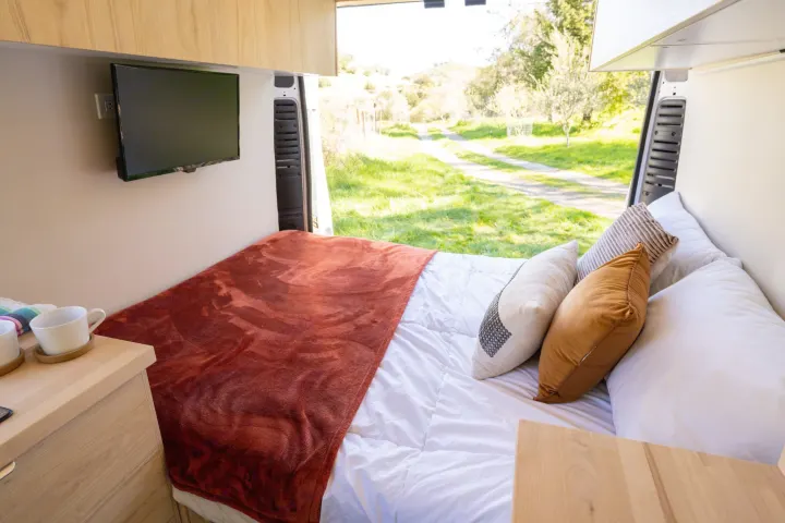 Comfortable RV rental bed setup with TV and open view to a scenic outdoor path.. ram ProMaster 2021