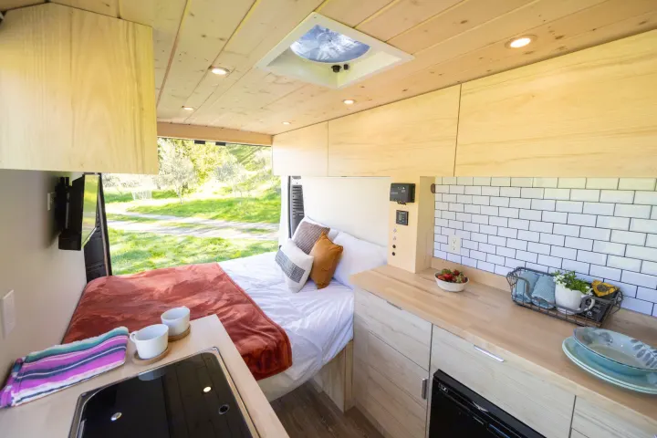 This rv rental offers a cozy, modern wood-finished interior with a comfortable bed, functional kitchen, and scenic outdoor views for van life.. ram ProMaster 2021