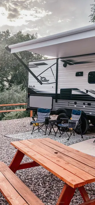 Relaxing at an RV rental campsite. Jay Flight Baja RV with extended awning, outdoor rug, chairs, and picnic table on gravel.. Jayco Featherlight SLX8 2022
