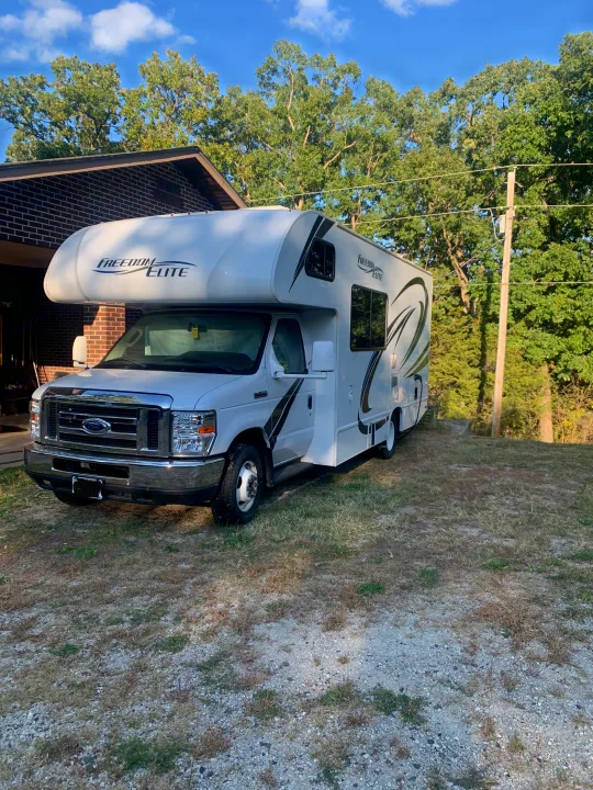 A white Freedom Elite Class C RV rental is parked on gravel next to a brick house, under a blue sky with trees.. Thor Freedom Elite 2019