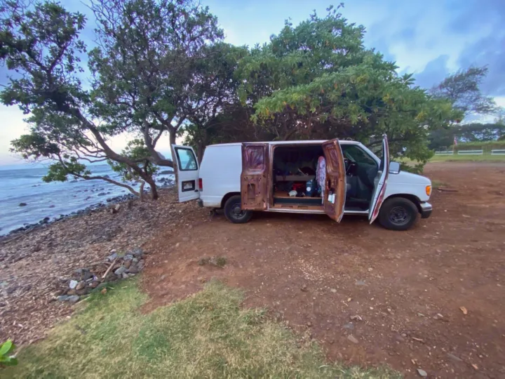 Oceanfront vanlife. A white converted van, doors open, parked by a rocky shore under trees. Escape in an RV rental alternative.. Ford Ford E150 Club Wagon 2003