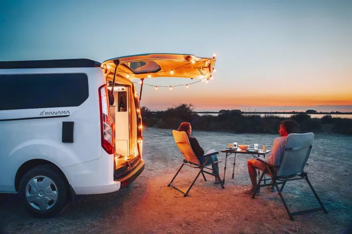 Relax at dusk with an RV rental. A camper van illuminates two people sitting at a table with string lights, enjoying the view.. Panama P10+ 2023