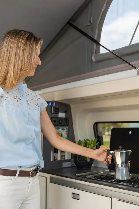 Start your day right in an RV rental! Woman makes coffee in a compact campervan kitchen with pop-top roof open.. Panama P10+ 2023