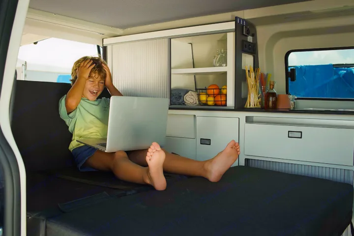 Child laughs on laptop in an RV rental, enjoying the compact, well-equipped interior with a kitchen setup and comfy bed.. Panama P10+ 2023
