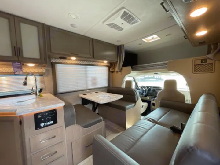 RV rental interior features a functional kitchen, dining booth, sofa, overhead bunk, and driver's cab view.. Thor Chateau 2019