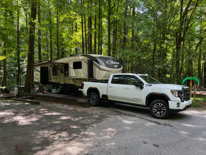 White GMC pickup truck and a large fifth-wheel RV parked at a wooded campsite, ideal for an rv rental adventure.. Keystone RV Cougar 2017